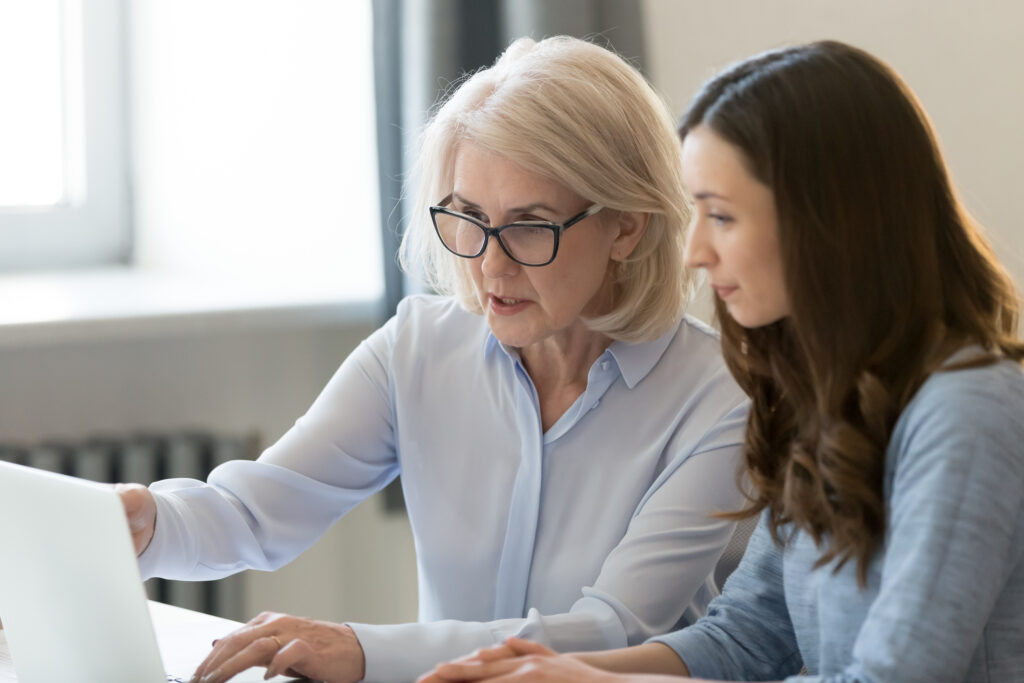 Older executive woman with black glasses and a light blue collared shirt points to a laptop screen as she teaches a younger woman new processes.