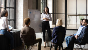 Confident lady standing by a whiteboard and speaking to seated coworkers